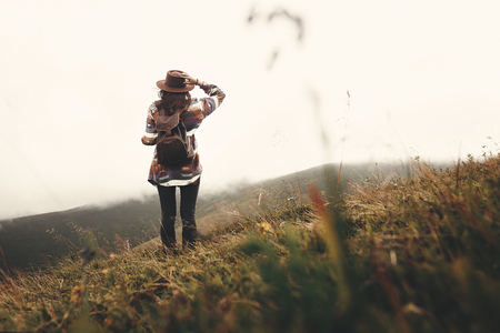 Stylish hipster girl in hat walking on top of mountains. Happy young woman with backpack exploring misty mountains. Travel and wanderlust concept. Amazing atmospheric momentの写真素材