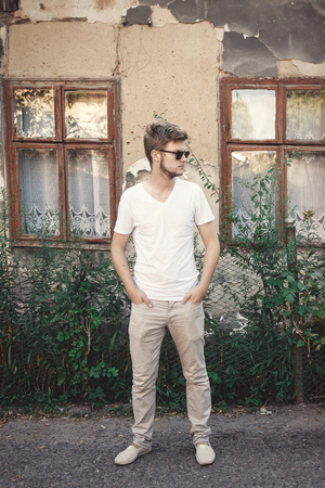 Stylish hipster man in sunglasses posing outdoors in sunny countryside at old rustic house. Handsome bearded man relaxing in evening summer city. Vacation and travelの写真素材