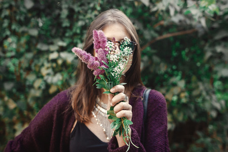 Portrait of happy boho girl in sunglasses smiling with bouquet of wildflowers in sunny garden. Stylish hipster carefree girl having fun at green bush in street. Summer vacation and travelの写真素材