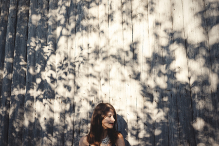 Stylish hipster girl posing in sunny street on background of wooden wall. Boho girl in cool outfit...