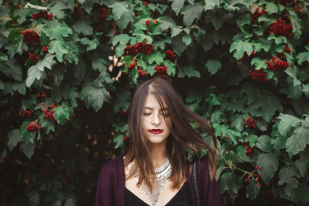 Stylish bohemian girl with modern jewelry posing under green branches of guelder rose in street. Hipster girl calm portrait among leaves and berries. Eco model. Summer vacationの写真素材