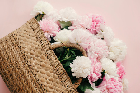Hello spring concept. Stylish straw rustic bag with white and pink peonies on pastel pink paper, flat lay with copy space. International Women's Day. Happy mothers day, floral greeting cardの写真素材