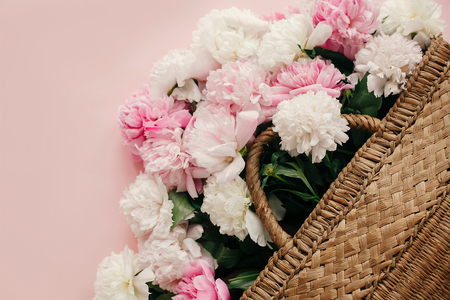 Stylish straw rustic bag with white and pink peonies on pastel pink paper, flat lay. Hello spring, creative image with copy space. Summer vacation concept. International Women's Day.の写真素材