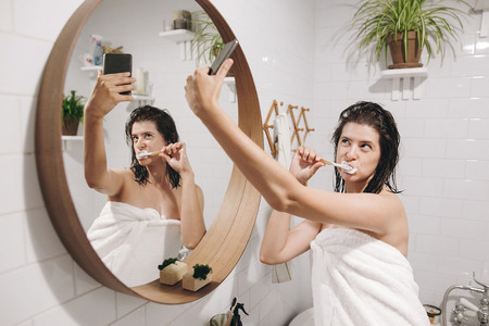 Young happy woman in white towel making selfie while brushing teeth and looking at phone in stylish bathroom at round mirror. Social media affect. Tooth care conceptの写真素材