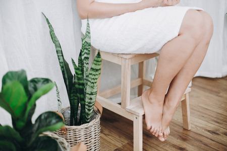 Young woman in white towel sitting in bathroom with green plants. Legs soft skin after shaving. Skin care and wellness concept.の写真素材