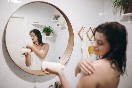 Young happy woman in white towel applying moisturizing cream on shoulder in bathroom, reflection in mirror. Skin and body care. Hand holding plastic bottle with lotion. Sexy woman relaxingの写真素材