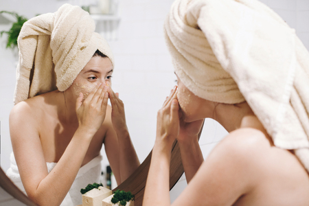 Young happy woman in towel applying organic face mask and looking at round mirror in stylish bathroom. Girl making facial massage with scrub, peeling and cleaning skin on face. Skin Careの写真素材