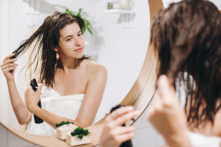 Hair and body care. Young happy woman in white towel applying conditioner mask on hair in bathroom, mirror reflection. Slim sexy woman with natural skin enjoying spa and wellness, relaxingの写真素材