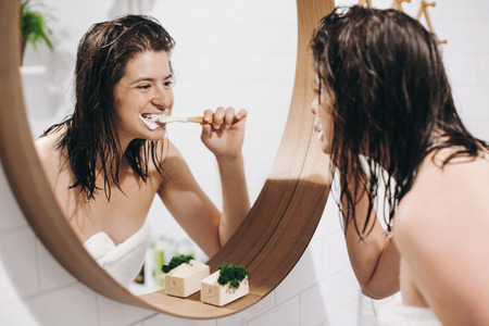 Tooth care concept. Young happy woman in white towel brushing teeth and looking at round mirror in stylish bathroom. Slim sexy woman daily routine after shower.の写真素材
