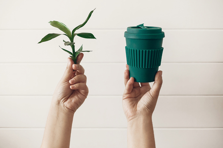 Zero waste concept, sustainable lifestyle. Hands holding stylish reusable eco coffee cup and green bamboo leaves on white wooden background. Ban single use plastic. Make a choice.の写真素材