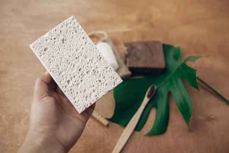 Hand holding natural eco sponge on background of bamboo toothbrush, soap, solid shampoo, crystal deodorant, on wood with green monstera leaf. Zero waste concept. Sustainable lifestyleの写真素材