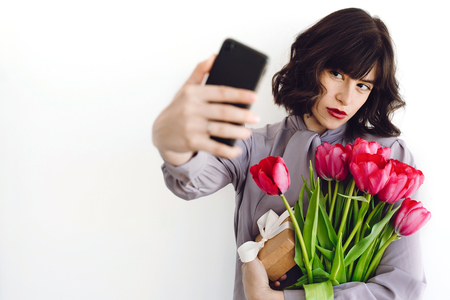 Beautiful brunette girl holding bouquet of tulips and gift box and taking selfie on white background indoors, space for text. Stylish young woman with flowers and phone. Happy mothers dayの写真素材