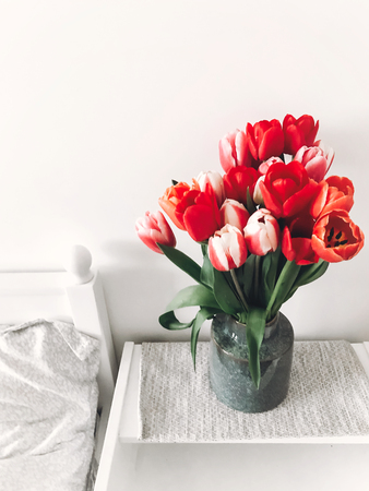 Beautiful red and pink tulips in vase on white wooden nightstand near stylish white bed, copy space. Hello spring concept. Happy mothers dayの写真素材