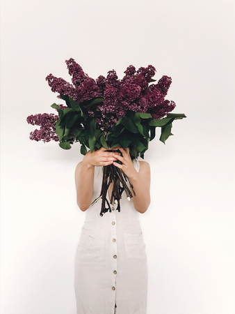Girl in rustic linen dress holding purple lilac flowers in front of face on white background. Hello spring. Happy Mothers day. Lilac bouquet in woman hands. International womens dayの写真素材
