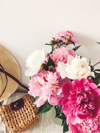 Stylish pink and white peonies in vase and trendy straw bag and hat on stylish white nightstand near bed. Hello spring. Happy Mothers day. Girly imageの写真素材