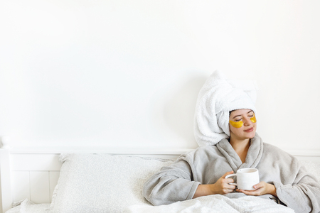 Beautiful young woman with eye patches in bathrobe holding cup of coffee and lying in bed, enjoying morning. Happy girl drinking coffee and applying eye patches, skin care and relax. Copy spaceの写真素材