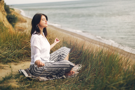 Young beautiful woman relaxing and practicing yoga on beach, sitting in grass and sand, meditating and listening to sea waves. Hipster girl doing yoga. Meditation,calm and happiness. Yoga dayの写真素材