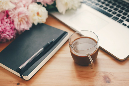 Stylish laptop, coffee cup, notebook, pen,  pink and white peonies on wooden table with space for...