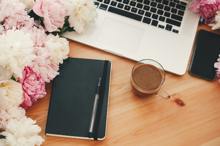Stylish laptop, coffee cup, notebook, pen,  pink and white peonies on wooden table with space for text. Freelance concept. International womens day. Stylish working place.の写真素材