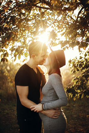 Happy young parents, mom and dad, hugging baby bump, enjoying beautiful moment at sunset. Stylish pregnant couple holding hands on belly and embracing in sunny light in autumn park under treeの写真素材