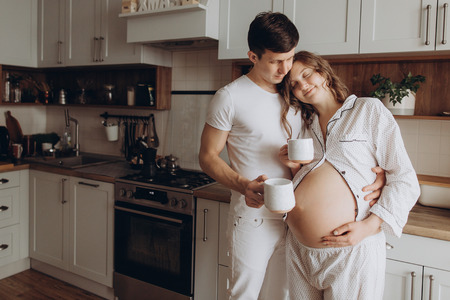 Happy young pregnant couple in white pajamas holding belly bump in kitchen in the morning. Stylish pregnant family, mom and dad relaxing at home, waiting for baby. Parenthood. Cute momentsの写真素材