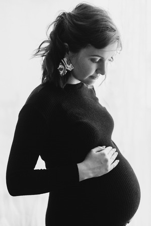 Happy young pregnant woman in stylish black dress holding belly bump and posing in light. Stylish...