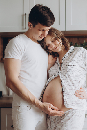 Happy young pregnant couple in white pajamas holding belly bump in kitchen in the morning. Stylish pregnant family, mom and dad relaxing at home, waiting for baby. Parenthood. Cute momentsの写真素材