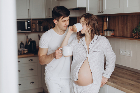 Happy young pregnant couple in white pajamas holding cups and relaxing in kitchen in the morning. Stylish pregnant family, mom and dad at home, waiting for baby. Cute moments of happiness.の写真素材