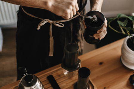 Professional barista in black stylish apron grinding coffee for aeropress, alternative brewing method. Hands holding manual grinder and aeropress, scales, kettle on wooden table, top viewの写真素材