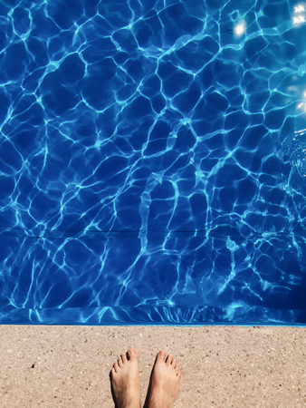 Tanned girl legs standing at pool with blue water in sunny light, top view. Summer vacation and relax. Space for text. Vertical phone photoの写真素材