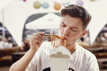 Stylish hipster man eating wok noodles with vegetables from carton box with bamboo chopsticks. Asian Street food festival. Guy tasting and eating delicious thai noodles in takeaway paper boxの写真素材