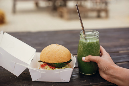Delicious vegan burger in carton tray and healthy spinach smoothie in glass jar with metal reusable straw  on wooden table at street food festival. Zero waste conceptの写真素材