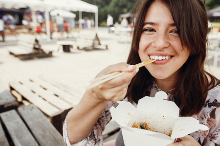 Stylish hipster girl eating wok noodles with vegetables from carton box with bamboo chopsticks. Asian Street food festival. Happy boho woman eating noodles in takeaway paper boxの写真素材