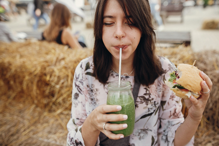 Stylish hipster girl holding delicious vegan burger and drinking smoothie in glass jar at street food festival. Happy boho woman tasting burger with healthy drink in summer street.の写真素材