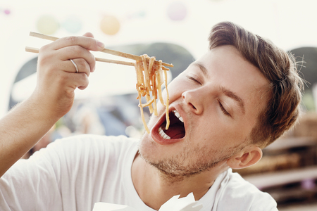 Stylish hungry man eating delicious wok noodles with vegetables from carton box with bamboo chopsticks. Asian Street food festival. Hipster guy tasting thai noodles in takeaway paper boxの写真素材