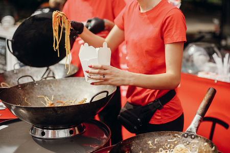 Chef putting noodles in carton box to go from pan on fire at open kitchen. Fried chinese japanese noodles with vegetables and shrimps in a wok on the open fire. Asian street food festivalの写真素材