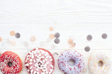 Delicious colorful donuts with sprinkles and marshmallows on stylish white table with confetti, flat lay, copy space. Party concept. No diet. Candy bar at wedding reception. Purple, pink donutsの写真素材