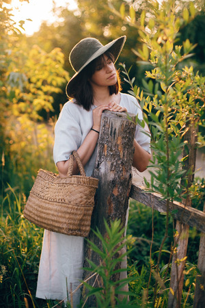 Stylish girl in linen dress holding rustic straw basket at wooden fence  in sunset light. Boho woman relaxing and posing in summer countryside in warm evening. Atmospheric rural momentの写真素材