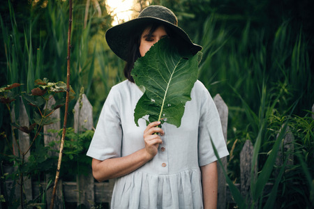 Stylish girl in linen dress holding big green leaf at face at wooden fence and grass. Portrait of boho woman in hat posing with leaf in summer countryside in evening. Atmospheric momentの写真素材