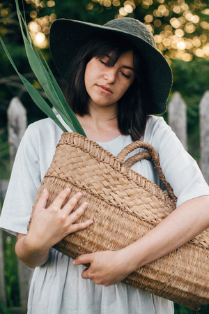 Stylish girl in linen dress holding rustic straw basket with green leaf at wooden fence and grass. Portrait of boho woman in hat posing in summer countryside in evening. Atmospheric momentの写真素材