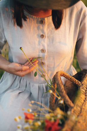 Stylish girl in linen dress gathering flowers in rustic straw basket, sitting in poppy meadow in sunset. Boho woman holding wildflowers bud in warm sunlight in summer field.の写真素材