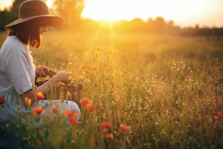 Stylish girl in linen dress gathering flowers in rustic straw basket, sitting in poppy meadow in sunset. Boho woman in hat relaxing in warm evening sunlight in summer field. Space for textの写真素材