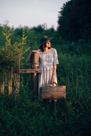 Stylish girl in linen dress holding rustic straw basket at wooden fence  in sunset light. Boho woman relaxing and gathering wildflowers in summer countryside. Atmospheric rural momentの写真素材