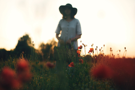 Focus on poppy flowers, blurred image of stylish girl in linen dress holding flowers in rustic straw basket in meadow in sunset light. Boho woman relaxing in summer field. Atmospheric momentの写真素材