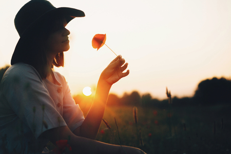 Silhouette of stylish girl in linen dress holding poppy flower in meadow in sun light with flowers in rustic straw basket. Boho woman in hat relaxing in summer sunset field. Atmosphericの写真素材
