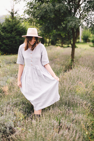 Stylish hipster girl in linen dress and hat walking in lavender field and relaxing. Happy bohemian woman enjoying lavender aroma in summer mountains. Atmospheric calm rural momentの写真素材