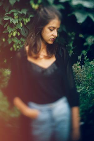 Stylish hipster girl posing at hop bush, atmospheric moment. Fashionable woman in denim jeans and black shirt relaxing in park. Selective focus, creative imageの写真素材