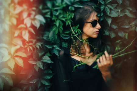 Stylish hipster girl posing at green leaves, atmospheric moment. Portrait of fashionable cool woman in black sunglasses relaxing in park. Selective focus, creative retro image. Space for textの写真素材