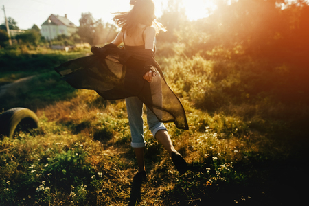 Stylish hipster girl having fun in sunny park in amazing sunbeams, atmospheric moment. Fashionable cool woman dancing in evening light, back view. Selective focus. Retro effect. Creative imageの写真素材