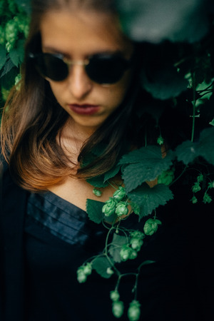 Stylish hipster girl posing at green hop bush, atmospheric moment. Portrait of fashionable cool woman in black sunglasses relaxing in park. Selective focus, creative image. Space for textの写真素材